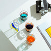 A collection of colorful, mouth-blown glasses with bold, graphic lines, displayed on a white table.