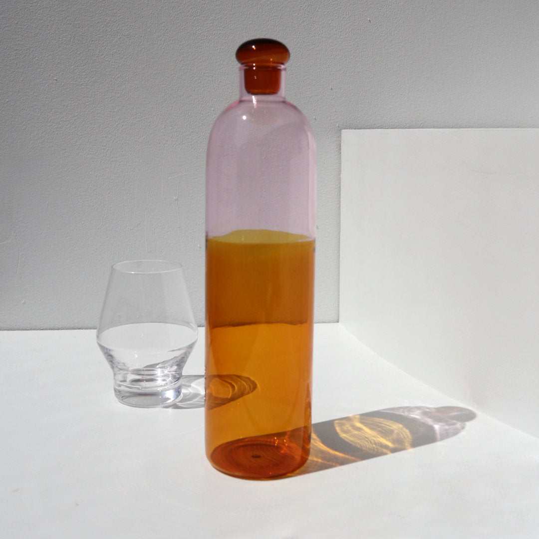 amber and pink bottle, with a glass bottle stop in bright amber shade, photographed in the direct sunlight on white background with a simple clear water glass 